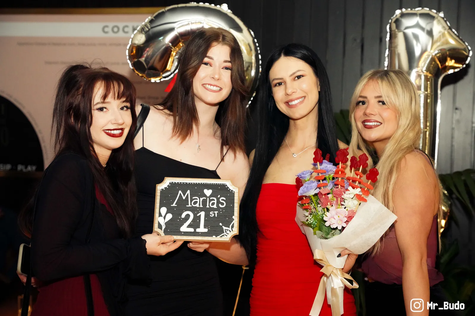 Four friends in black and red dresses celebrating a 21st birthday with a chalkboard sign and bouquet at a Canberra venue