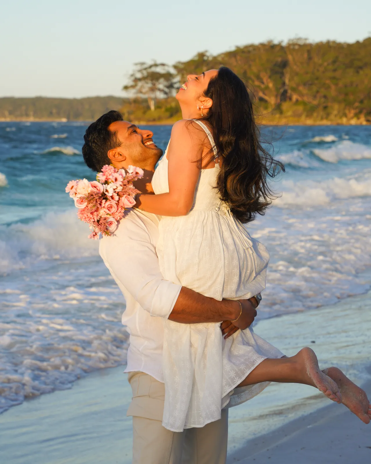 Groom lifting his laughing bride holding a bouquet in the surf during a beach wedding shoot