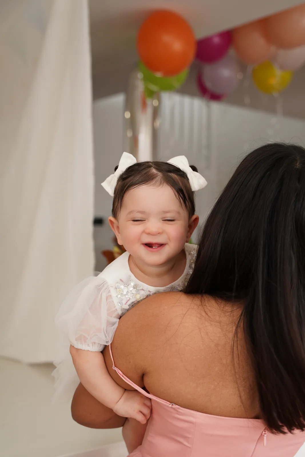 Smiling toddler with a hair bow over her mother's shoulder at a kids birthday party with balloons