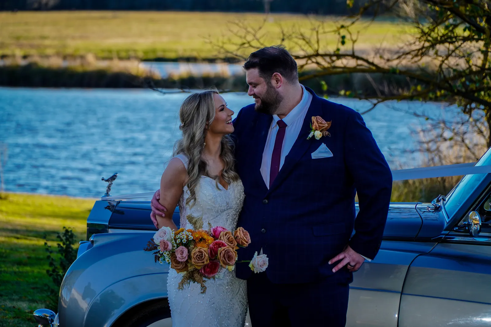 Bride and groom smiling at each other by a vintage car beside a lake at a Canberra wedding