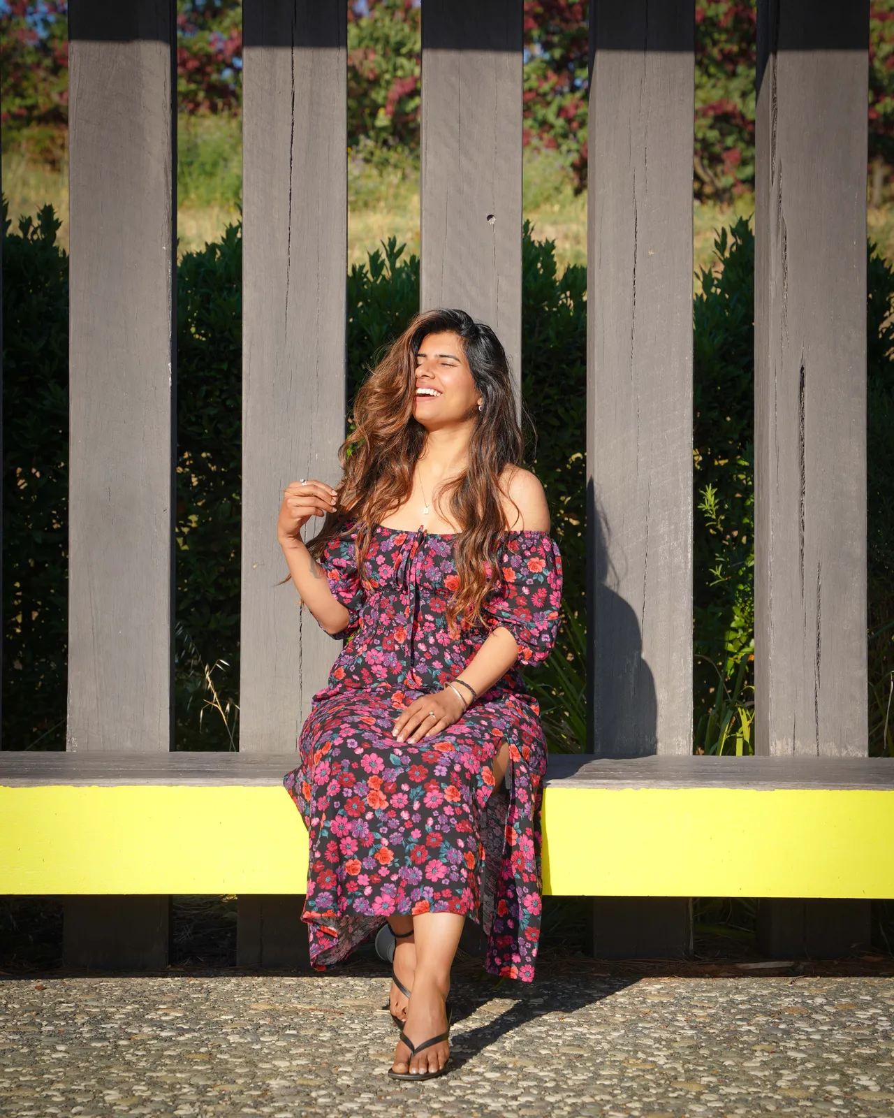 Woman in a floral dress laughing on a yellow bench against a timber fence in an outdoor portrait